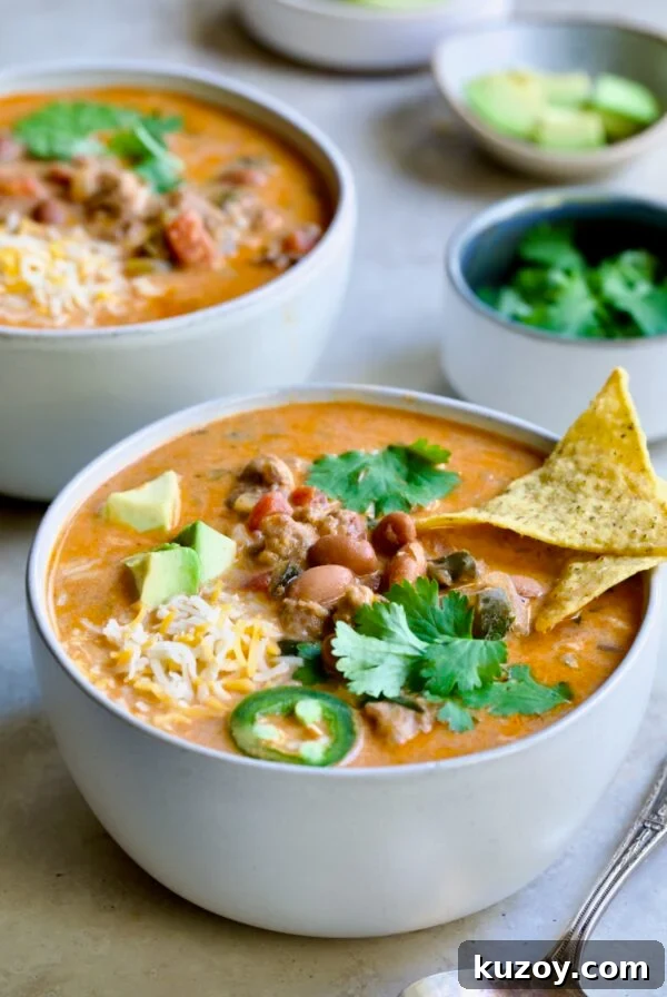One serving of Creamy Taco Soup in white bowl garnished with tortilla chips, avocado, cheese and jalapeno slices.
