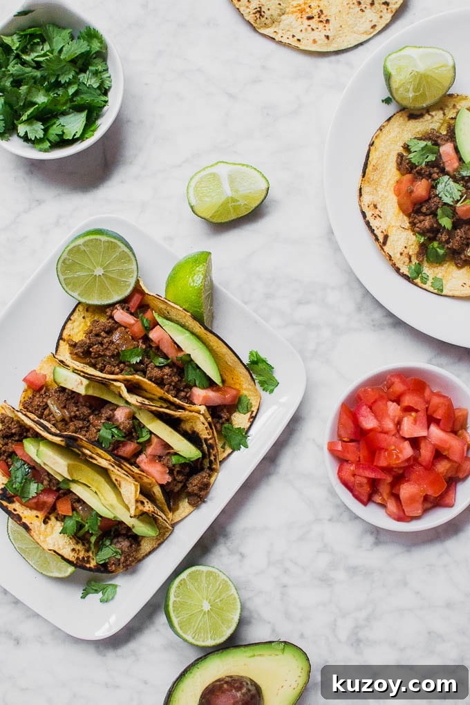 Gluten-Free Beef Tacos: Flavorful Corn Tortilla Recipe 4 Aerial view of a plate of gluten-free ground beef tacos, with a bowl of diced tomatoes, a half of an avocado, a bowl of cilantro, and limes.