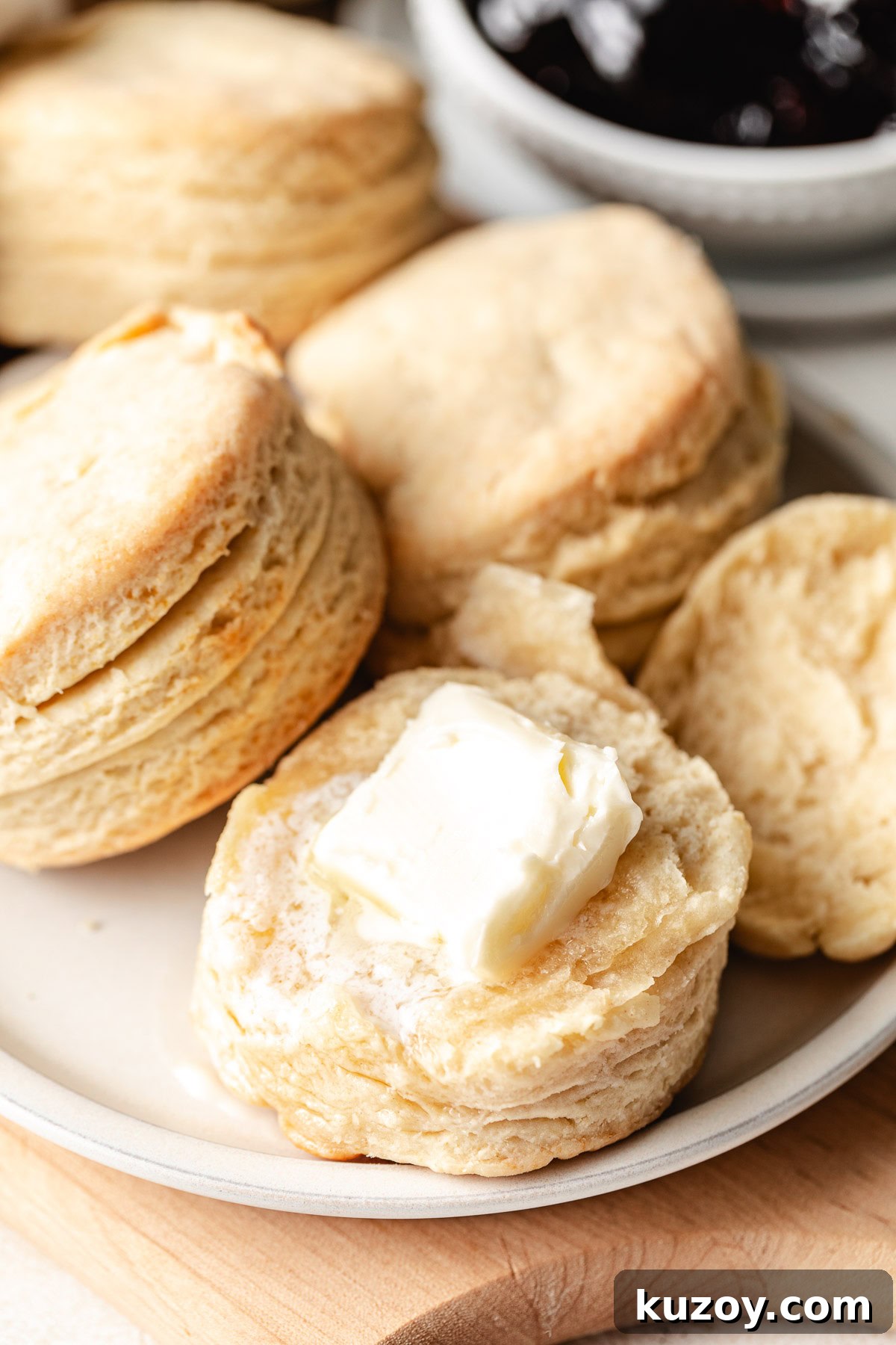 Plate of homemade buttermilk biscuits with a pat of butter on top and a bowl of jam in the background.