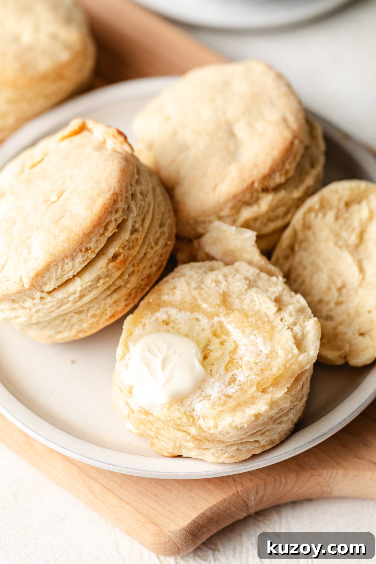 Close up of a flaky homemade buttermilk biscuit with butter on a white plate, showing layers on the side.