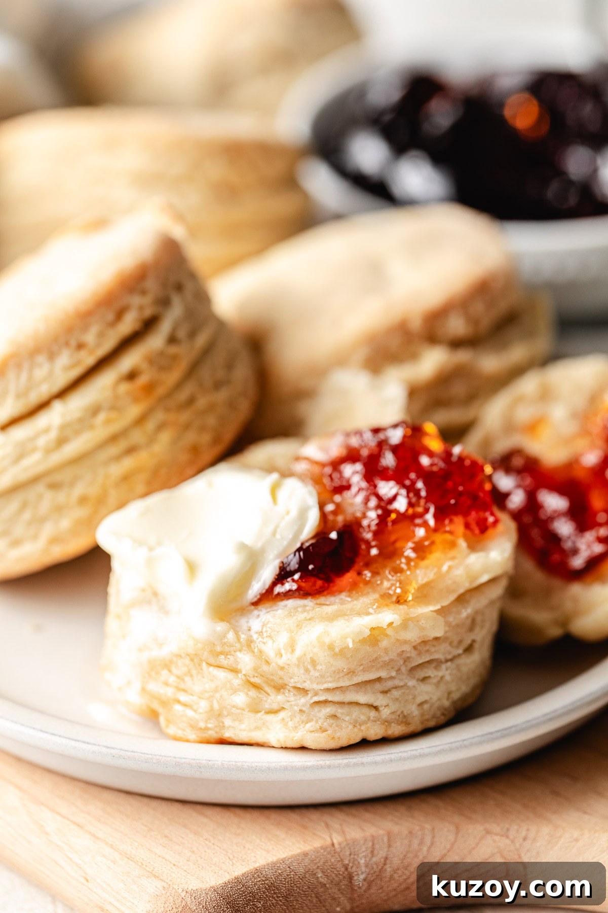 Close up of a flaky homemade biscuit with butter and strawberry jam, showing golden layers on the side.