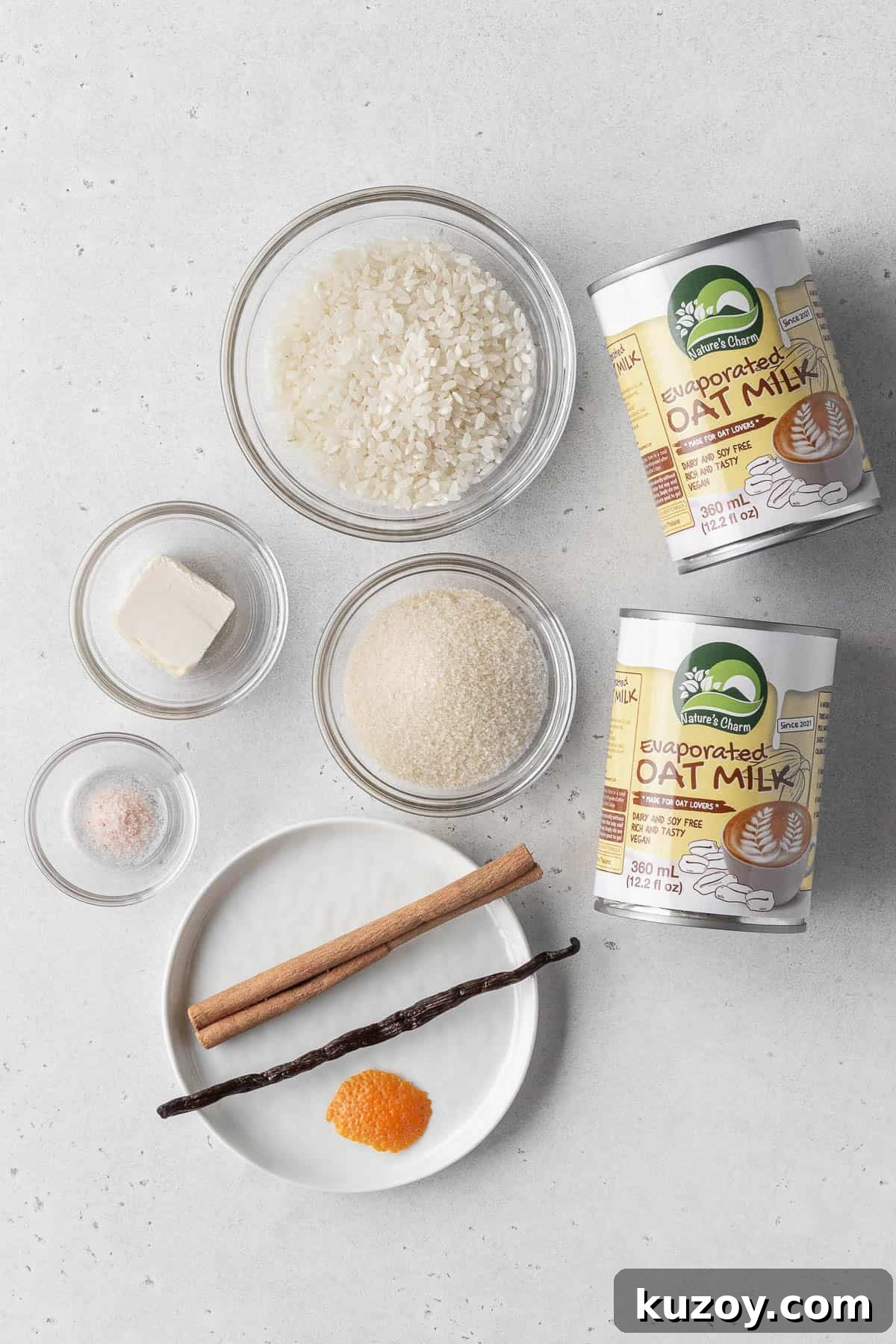 Creamy Dairy-Free Vegan Rice Pudding Recipe 3 Vegan rice pudding ingredients measured out into bowls with 2 cans of evaporated oat milk on a white surface.