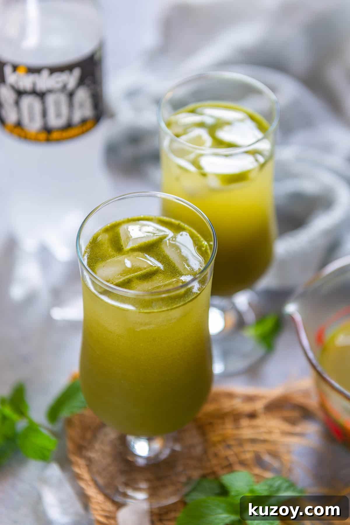 Two glasses filled with masala soda sit on a woven mat, with fresh mint leaves nearby. A partially visible bottle of soda water in the background while a glass jug in the background adds to the bright and refreshing scene.