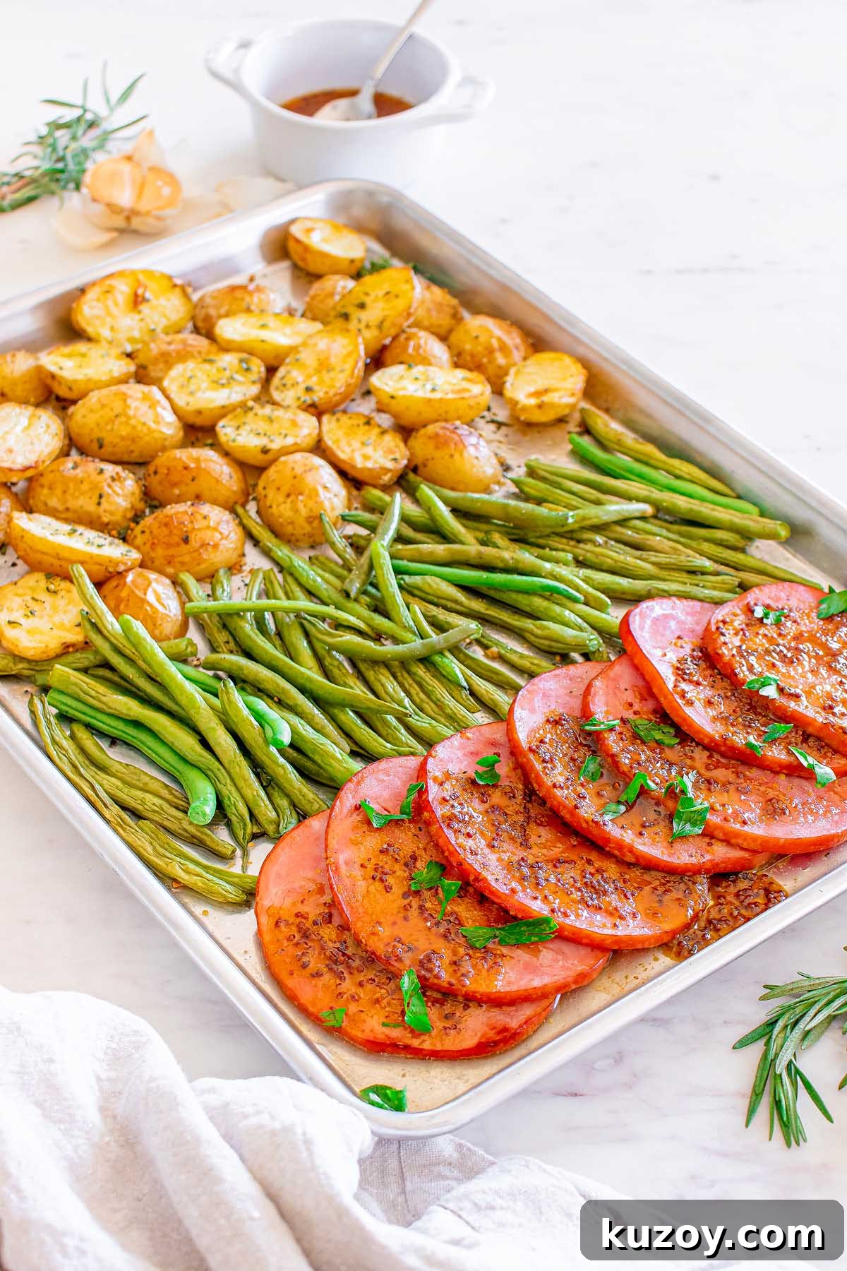 One-Pan Baked Ham Dinner with Roasted Vegetables 8 Baking sheet with sliced ham, green beans and halved potatoes, garnished with rosemary.