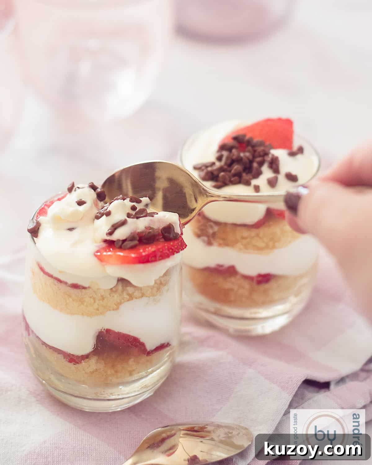 Two glasses filled with strawberry mascarpone cream and cake trifles. A golden spoon takes a bite.
