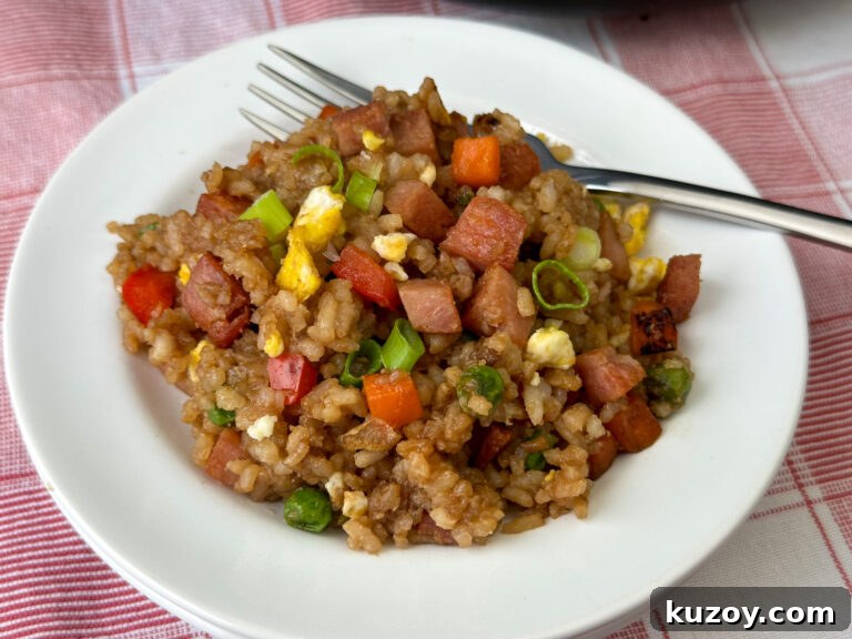 Chopped bits of SPAM resting on top of fried rice with pieces of egg, red pepper, carrots and slices of green onions on a plate resting on a red and white plaid napkin.