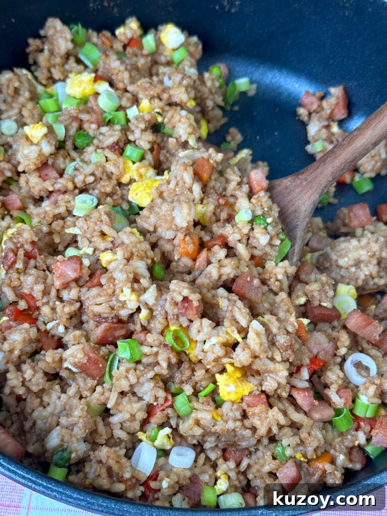 Close up fried rice with soy sauce, small pieces of vegetables and spam with a wooden spoon about to lift up a serving.