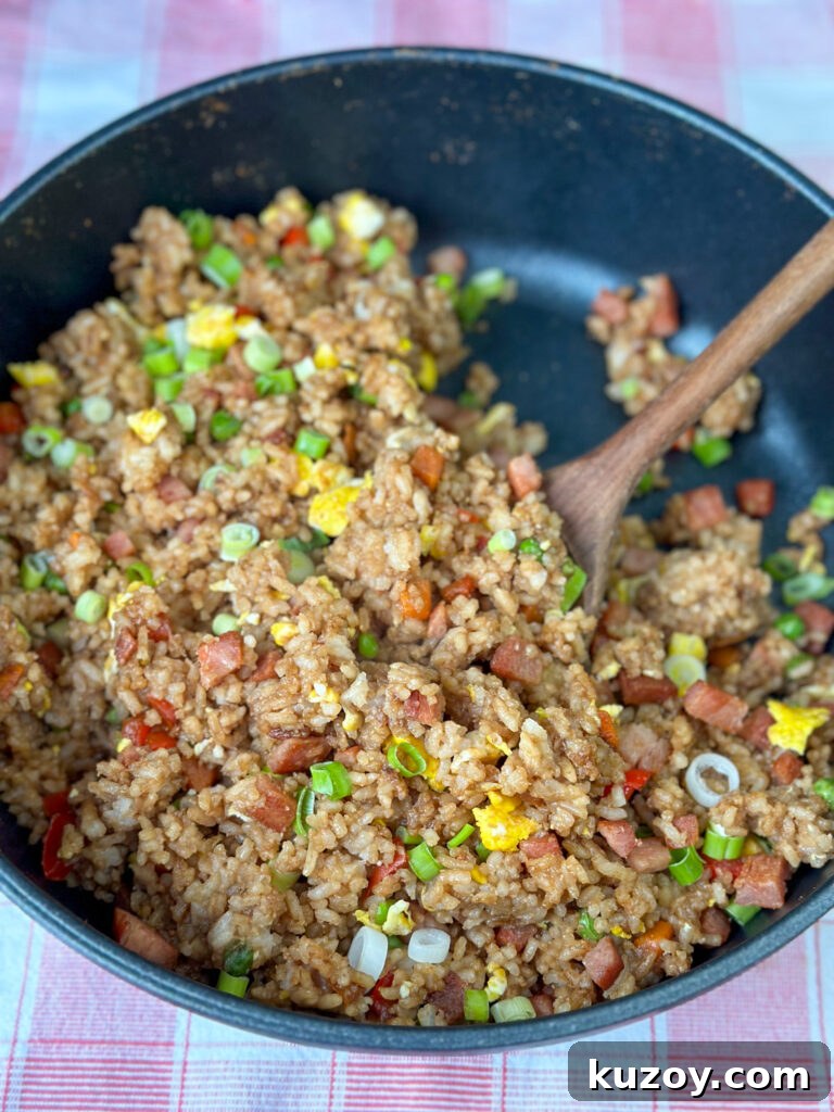 Brown wooden spoon stirring spam fried rice in a deep non-stick skillet.